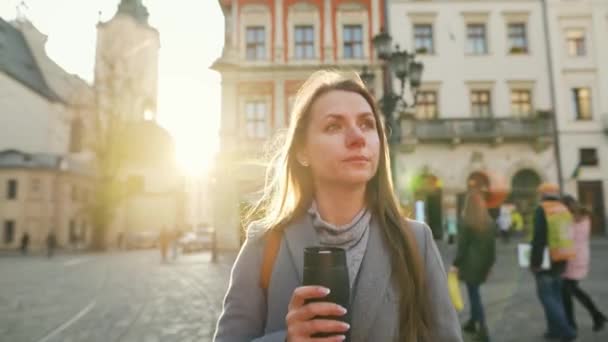 Mulher com uma xícara térmica na mão andando pela rua e admira a arquitetura da cidade velha ao pôr do sol em um dia frio de outono — Vídeo de Stock