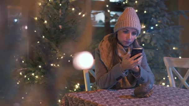 女性は冬に屋外のカフェテラスに座って、背景にクリスマスの装飾が施されたスマートフォンを使用してソーシャルネットワークでチャットします。 — ストック動画