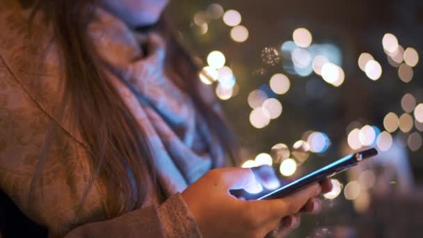 Primer plano de la mujer utiliza un teléfono inteligente en invierno en el fondo de un árbol de Navidad decorado con luces. Comunicación, Navidad y Año Nuevo Concepto de vacaciones — Vídeo de stock