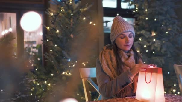 Frau sitzt im Winter auf einer Café-Außenterrasse und schaut in eine Tüte mit einem Geschenk und freut sich über das, was sie dort findet. Das Paket wird von innen hervorgehoben — Stockvideo