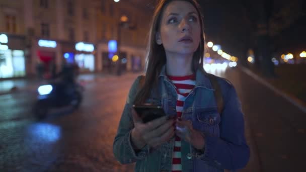 Vrouw met smartphone en overweegt de omgeving tijdens het wandelen door de straten van de avondstad — Stockvideo