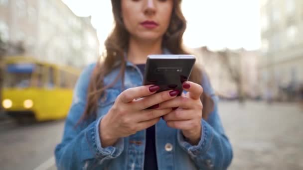 日没時にスマートフォンを使って古い通りを歩いている女性。黄色のトラムが背景に乗っています。コミュニケーション、ソーシャルネットワーク、オンラインショッピングの概念 — ストック動画