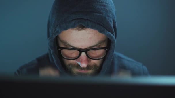 フードを着た男性ハッカーと暗いオフィスの部屋でコンピュータに取り組んでいる眼鏡。サイバー犯罪の概念 — ストック動画
