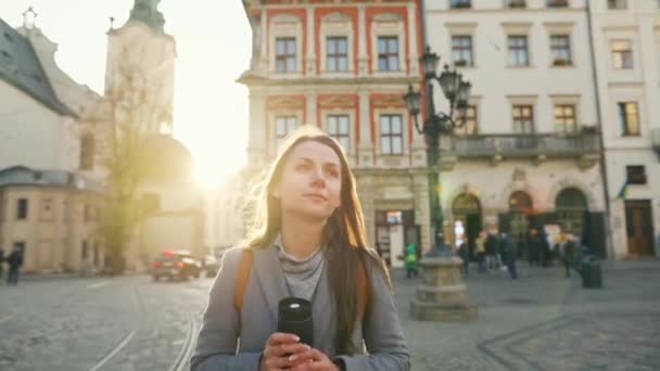 Жінка з чашкою термос в руці йде по вулиці і захоплюється архітектурою старого міста на заході сонця в холодний осінній день — стокове відео