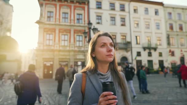 Kvinna med en termos kopp i handen står mitt på gatan och tittar på hennes klocka, väntar på den som är sen att träffa henne vid solnedgången på en kall höstdag — Stockvideo