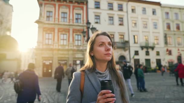 Mulher com uma taça térmica na mão fica no meio da rua e olha para seu relógio, esperando por aquele que está atrasado para encontrá-la ao pôr do sol em um dia frio de outono — Vídeo de Stock