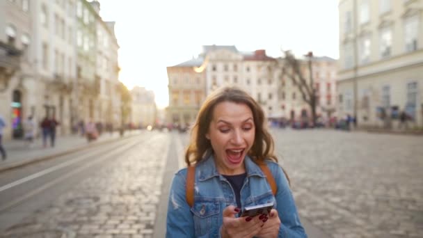 Eski bir caddede akıllı telefon kullanarak yürüyen bir kadın orada gördüğü için çok mutlu. Kazanmak ya da büyük şans konsepti. Yavaş çekim — Stok video