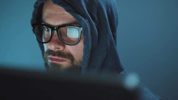 Un pirate dans le capot et des lunettes travaillant sur un ordinateur dans une pièce sombre du bureau. Concept de cybercriminalité — Video