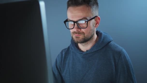 Bearded male hacker in hoodie and glasses working on a computer in a dark office room. Cybercrime concept — Stock Video