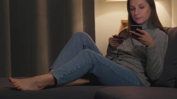 女人躺在舒适房间的沙发上，用信用卡和智能手机在网上购物。 网上购物、生活方式技术. — 图库视频影像
