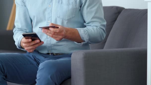 Formellt klädd man sitter på en soffa och skriver in ett kreditkortsnummer i en smartphone för att betala online. Onlineköp, livsstilsteknik. Närbild — Stockvideo