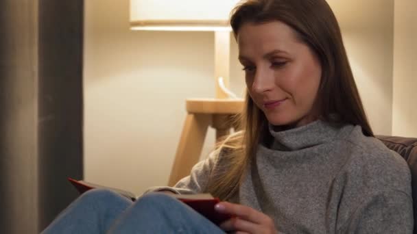 Женщина лежит на диване в уютной комнате и читает книгу вечером при свете лампы — стоковое видео