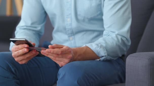 Un homme habillé formellement s'assoit sur un canapé et entre un numéro de carte de crédit dans un smartphone pour payer en ligne. Achats en ligne, technologie lifestyle. Gros plan — Video