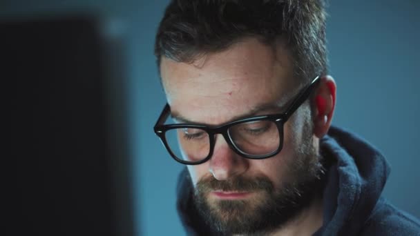 Бородатый мужчина-хакер в толстовке и очках, работающий за компьютером в темной офисной комнате. Концепция киберпреступлений — стоковое видео