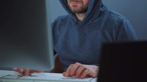 Un hacker maschio con cappuccio e occhiali che lavora su un computer in una stanza buia dell'ufficio. Concetto di criminalità informatica — Video Stock