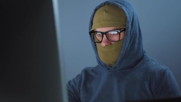 Ein männlicher Hacker mit Gesichtsmaske und Brille arbeitet in einem dunklen Büroraum an einem Computer. Cybercrime-Konzept — Stockvideo