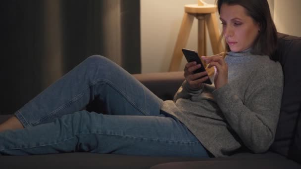 Usmívající se žena ležící na gauči v útulné místnosti a pomocí smartphone pro surfování na internetu ve večerních hodinách. Relaxační a životní styl technologie. — Stock video