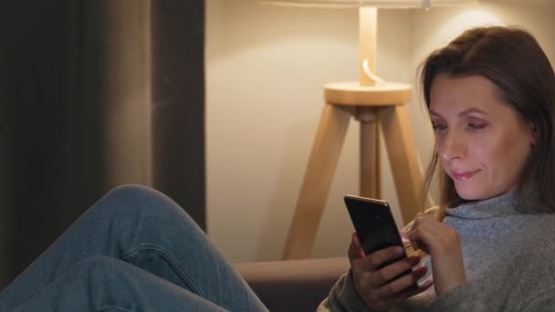 Mujer sonriente acostada en el sofá en una habitación acogedora y el uso de un teléfono inteligente para navegar por Internet por la noche. Tecnología de relajación y estilo de vida . — Vídeo de stock