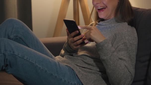 Sıcak bir odada kanepede uzanmış gülümseyen bir kadın internette sörf yapmak ya da akşamları biriyle sohbet etmek için akıllı telefon kullanıyor. Gördüğü için çok mutlu. — Stok video
