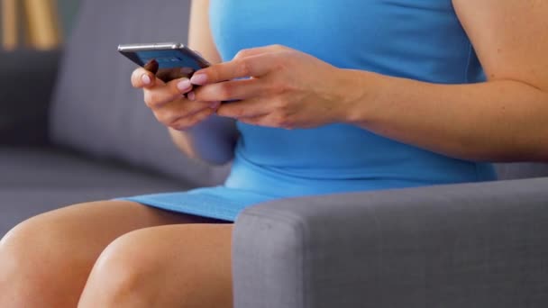 Femme en robe bleue s'assoit sur un canapé et utilise un smartphone pour envoyer un message texte. Gros plan — Video