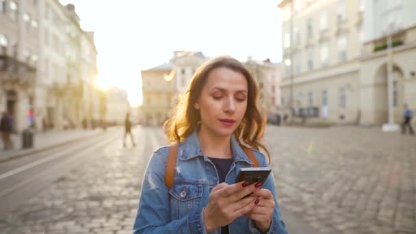 Mulher andando por uma rua velha usando smartphone ao pôr do sol. Comunicação, conceito de redes sociais. Movimento lento — Vídeo de Stock