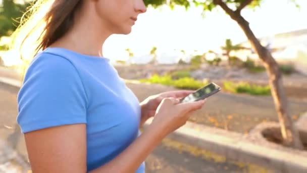 Vrouw in een blauwe jurk met smartphone terwijl ze op straat staat met een tropisch landschap — Stockvideo