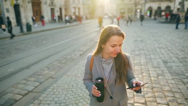Mujer con una taza de termo en la mano caminando por una calle vieja usando un teléfono inteligente al atardecer. Comunicación, redes sociales, concepto de compras en línea . — Vídeo de stock