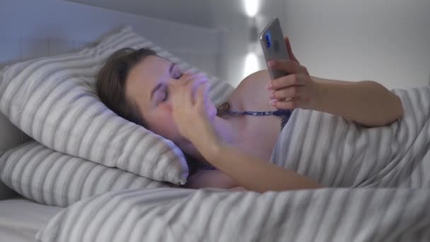 Femme utilise un smartphone alors qu'elle est couchée au lit. Elle se frotte les yeux, parce qu'elle est fatiguée et endormie. Concept de dépendance mobile ou d'insomnie . — Video