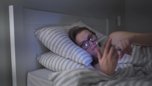 Mujer con gafas utiliza un teléfono inteligente mientras está acostado en la cama. Ella se queda dormida en el proceso porque está muy cansada — Vídeos de Stock