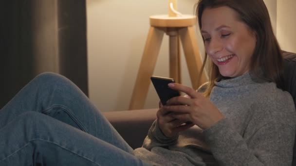 Lachende vrouw liggend op de bank in een gezellige kamer en met behulp van smartphone surfen op internet in de avond. Ontspanning en lifestyle technologie. — Stockvideo