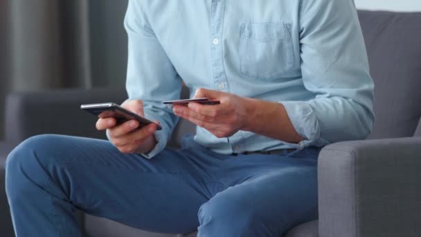 Un homme habillé formellement s'assoit sur un canapé et entre un numéro de carte de crédit dans un smartphone pour payer en ligne. Achats en ligne, technologie lifestyle. Gros plan — Video