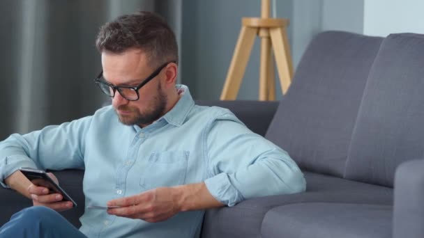 Homem barbudo de óculos senta-se no chão perto do sofá e faz o pagamento on-line com um cartão de crédito e smartphone. Compras online, loja no sofá, tecnologia de estilo de vida — Vídeo de Stock