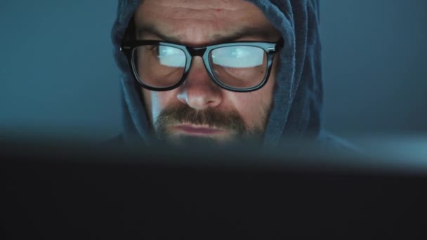 フードを着た男性ハッカーと暗いオフィスの部屋でコンピュータに取り組んでいる眼鏡。サイバー犯罪の概念 — ストック動画