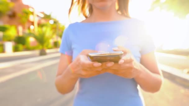 Frau in blauem Kleid mit Smartphone beim Spaziergang auf einer Palmenstraße bei Sonnenuntergang — Stockvideo