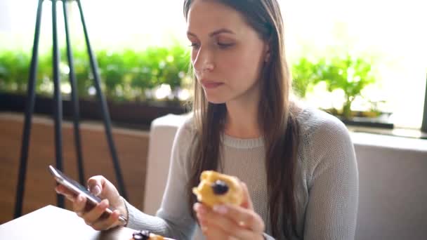 Kvinna äter eclairs i ett café och använda en smartphone — Stockvideo