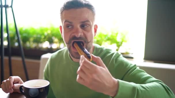 Homem barbudo com bigode enrolado come eclair de chocolate e bebe café no café — Vídeo de Stock