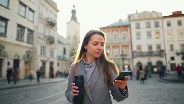 Egy nő egy termosz csészével a kezében, aki egy régi utcán sétál okostelefonnal naplementekor. Kommunikáció, közösségi hálózatok, online vásárlási koncepció. — Stock videók