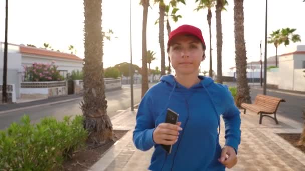 Kvinna med hörlurar och smartphone springer nerför gatan längs palmavenyn vid solnedgången. En hälsosam aktiv livsstil. Långsamma rörelser — Stockvideo