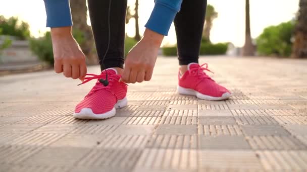 Жінка зав'язує шнурки на кросівки, вставляючи навушники в вуха і починає бігати по долоні на заході сонця — стокове відео