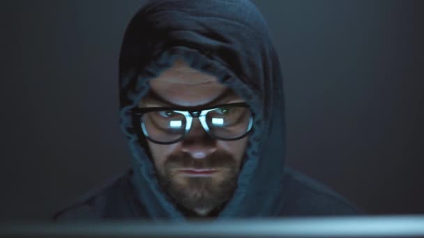 Ein männlicher Hacker unter der Haube und eine Brille, die in einem dunklen Büroraum an einem Computer arbeitet. Cybercrime-Konzept — Stockvideo