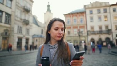 Elinde termos bardağı olan bir kadın gün batımında akıllı telefon kullanarak eski bir caddede yürüyor. İletişim, sosyal ağlar, online alışveriş kavramı.