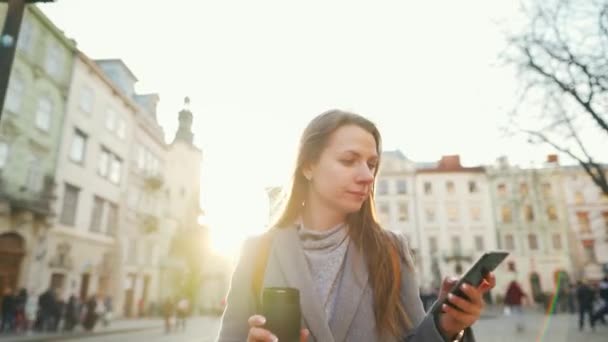 Mujer con una taza de termo en la mano caminando por una calle vieja usando un teléfono inteligente al atardecer. Comunicación, redes sociales, concepto de compras en línea . — Vídeo de stock