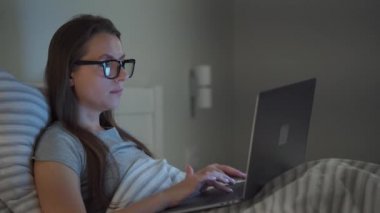 Woman in glasses working on laptop while lying in bed at night. Mobile addict or insomnia concept.