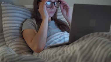 Woman in glasses working on laptop while lying in bed at night. She rubs her eyes, because she is tired and sleepy. Concept of increased stress and fatigue.