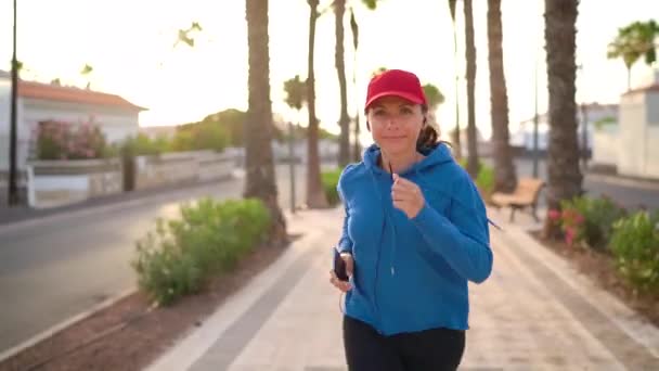 Woman with headphones and smartphone runs down the street along the palm avenue at sunset. Healthy active lifestyle — Stockvideo