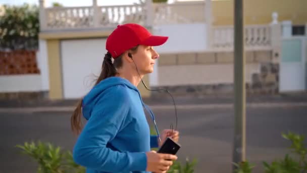 Mujer con auriculares y smartphone corre por la calle. Vida activa saludable. En cámara lenta. Vista lateral — Vídeos de Stock