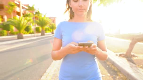 Woman in a blue dress using smartphone while walks on a palm street at sunset — 비디오