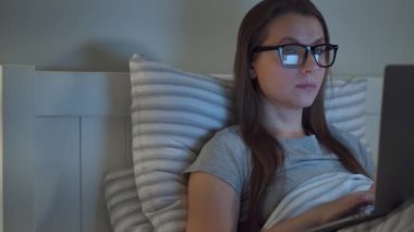 Woman in glasses working on laptop while lying in bed at night. She rubs her eyes, because she is tired and sleepy. Concept of increased stress and fatigue.