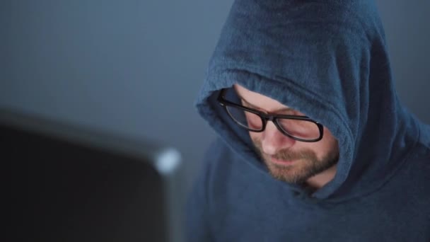 Un pirate dans le capot et des lunettes travaillant sur un ordinateur dans une pièce sombre du bureau. Concept de cybercriminalité — Video