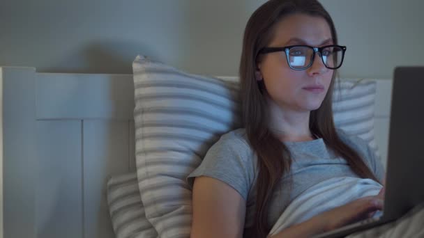 Женщина в очках работает на ноутбуке, лежа ночью в постели. Она потирает глаза, потому что устала и хочет спать. Концепция повышенного стресса и усталости . — стоковое видео